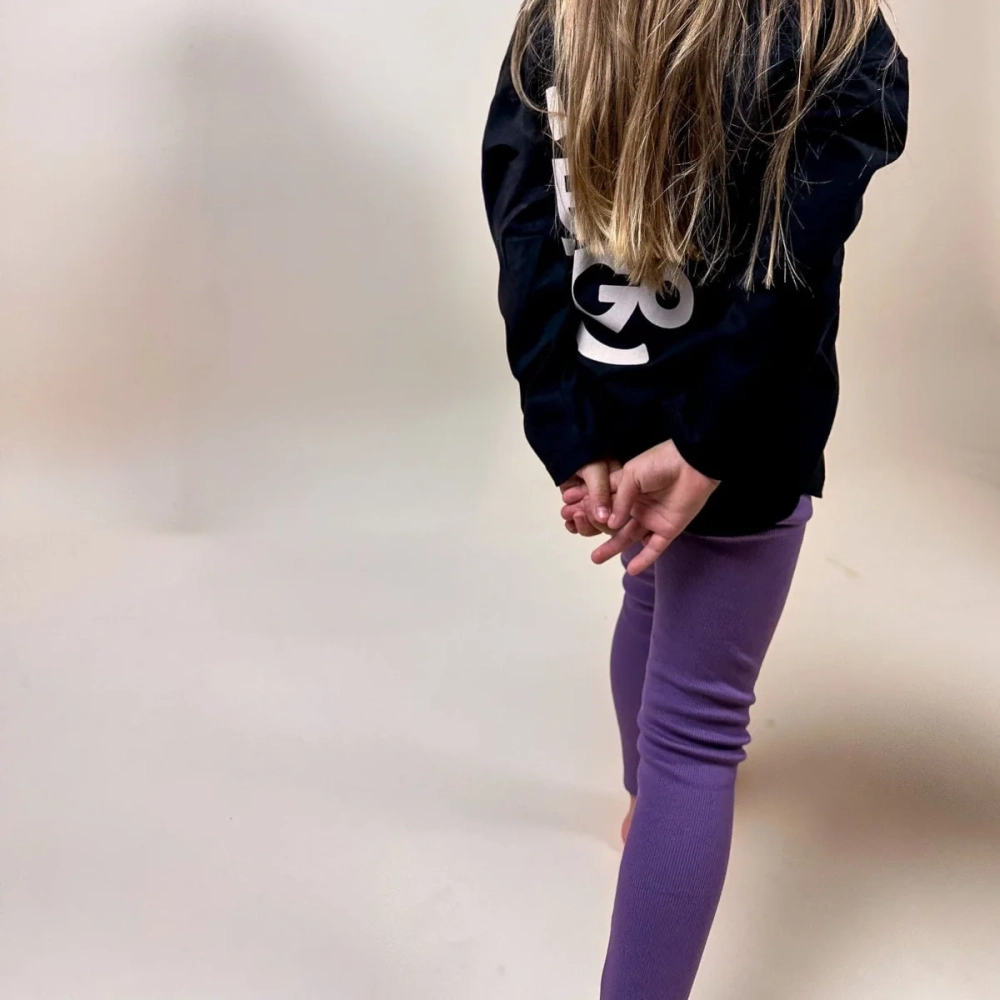 Dziewczynka stojąca tyłem, ubrana w fioletowe, jagodowe legginsy Fawn i czarną bluzkę z logo Buogo – wygodna i modna odzież dziecięca, idealna na zabawę i codzienne aktywności