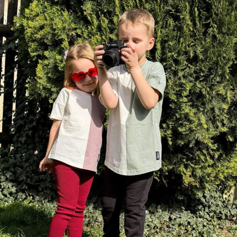 Chłopiec i dziewczynka na zewnątrz, ubrani w dwukolorowe t-shirty Moss marki Buogo: chłopiec w kremowo-miętowym, dziewczynka kremowo-jagodowym, korzystają z aparatu fotograficznego i mają okulary przeciwsłoneczne, które podkreślają ich styl.