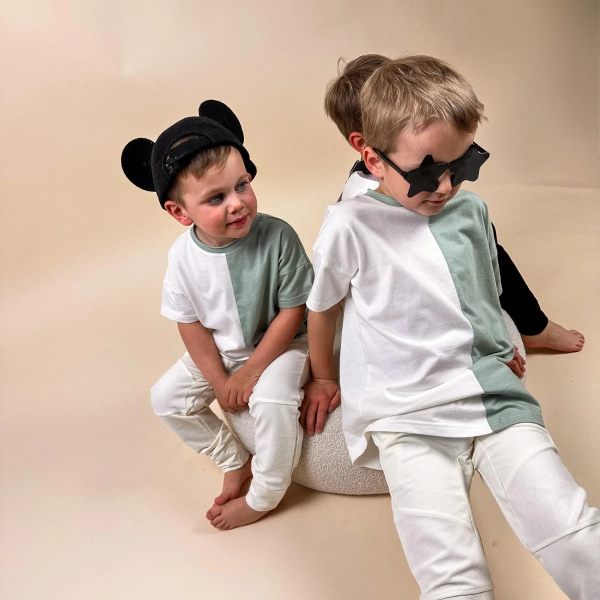 Dwoje dzieci w modnych dwukolorowych t-shirtach Moss marki Buogo - kremowo-miętowych, w jasnych spodniach, pokazują swoje urocze stylizacje w studio.