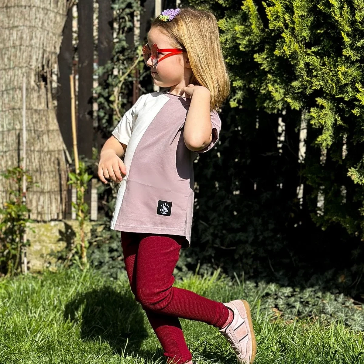 Mała dziewczynka biegająca po ogrodzie w stylowym asymetrycznym t-shircie Moss w odcieniach jagodowym i kremowym, połączonym z bordowymi (wiśniowymi) legginsami - modny i wygodny look.