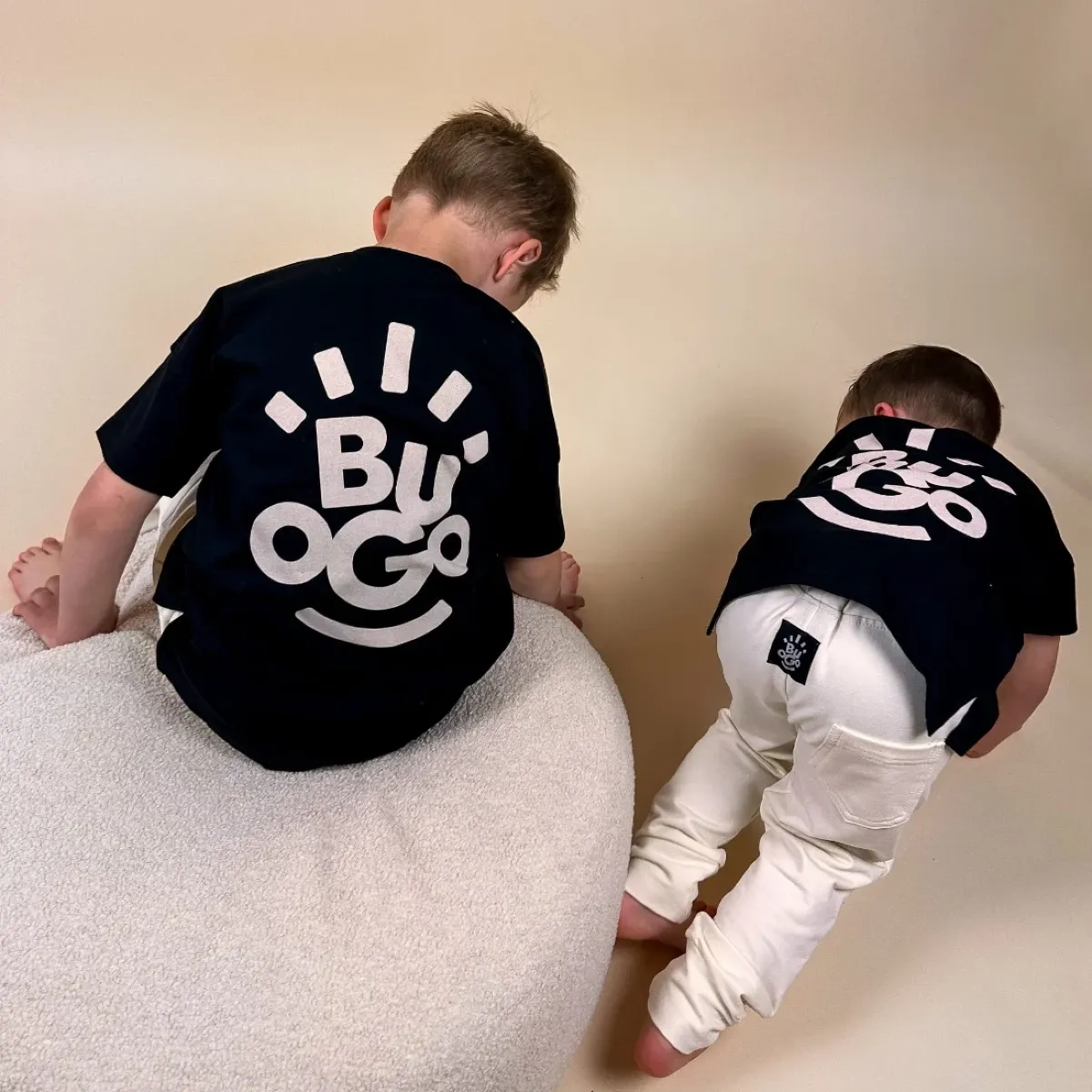 Dwóch chłopców siedzi na miękkiej pufie, ubrani w czarne koszulki Daylight z dużym białym logo Buogo na plecach oraz jasne spodnie – wygodna odzież dziecięca na co dzień.