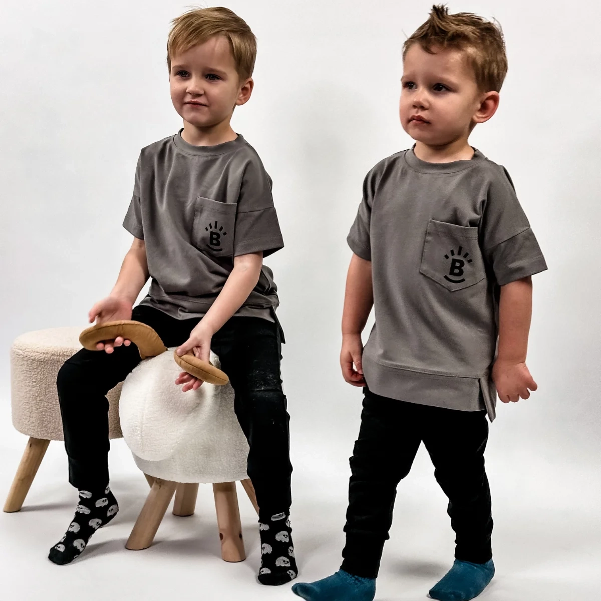 Dwaj uśmiechnięci chłopcy w szarych koszulkach Deer marki Buogo, jeden siedzi na pufie, drugi stoi obok niego - doskonały wybór na codzienne zabawy i wspólne chwile.