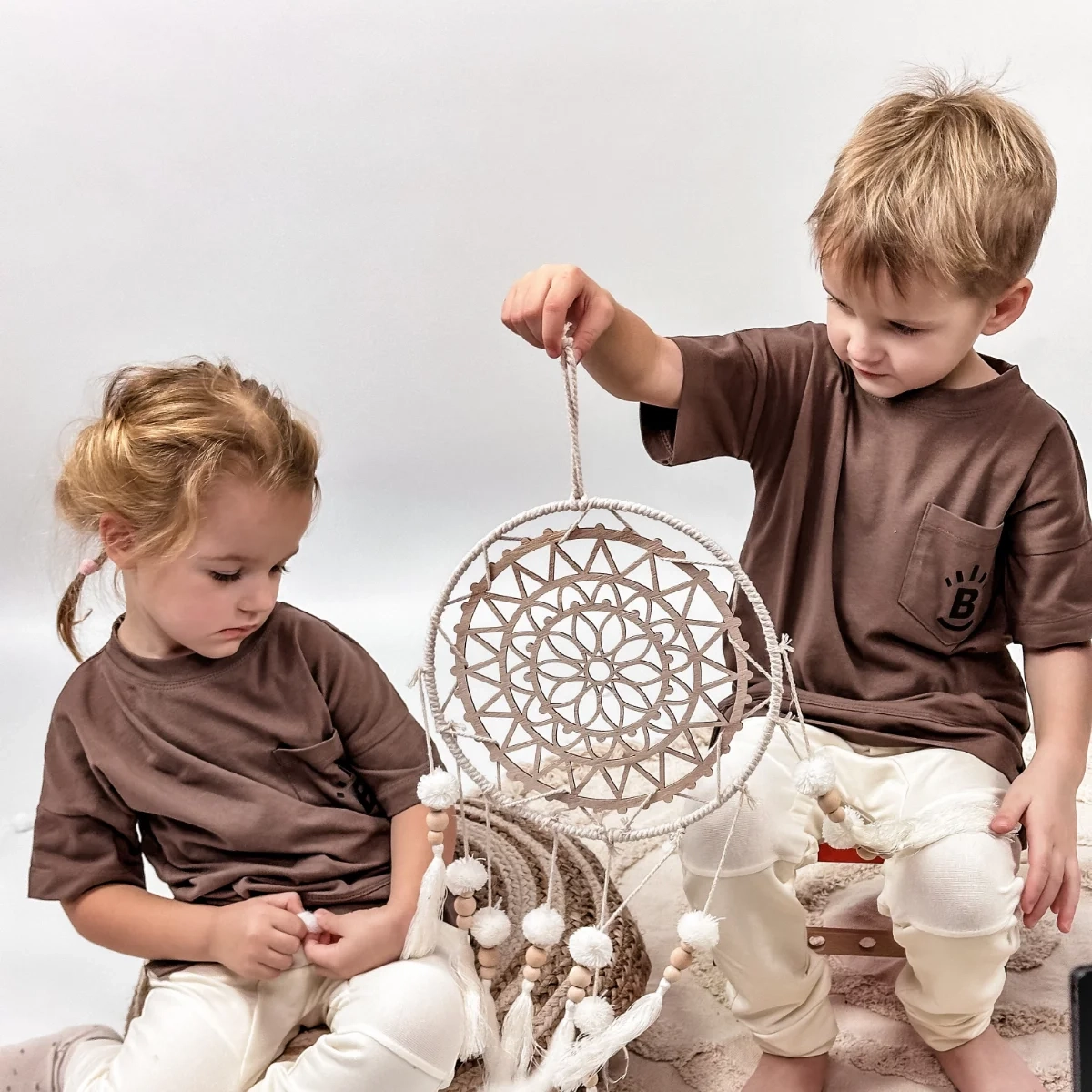 Dziewczynka z chłopcem bawią się razem, trzymają łąpacz snów, ubrani w brązowe (kakaowe)  koszulki, T-shirty Deer marki Buogo - wygoda i styl na co dzień.